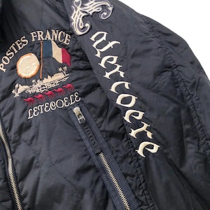 Puede incluir: Chaqueta acolchada azul marino con detalles bordados. Presenta una bandera francesa, las palabras "Postes France" y "Leteocole" con un dise&ntilde;o de camellos. La manga derecha tiene la palabra "Uercoere" en blanco.