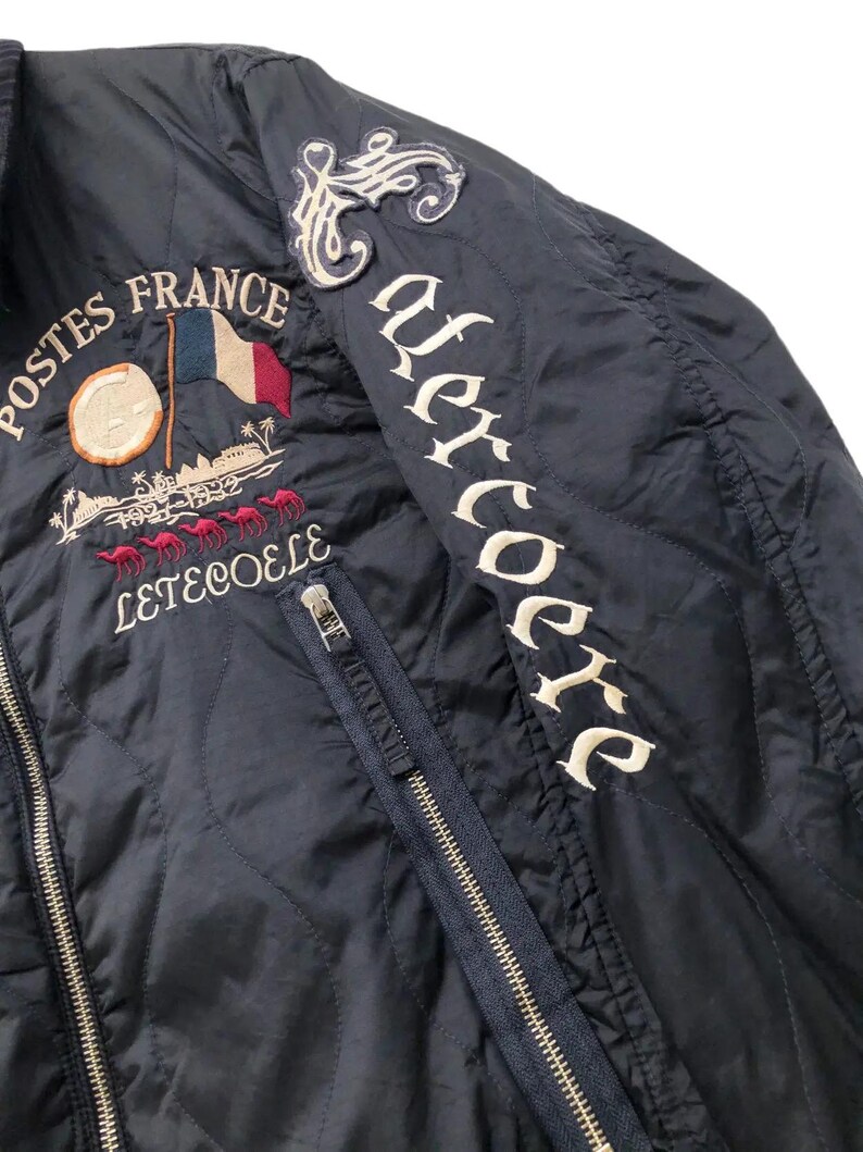 Puede incluir: Chaqueta acolchada azul marino con detalles bordados. Presenta una bandera francesa, las palabras "Postes France" y "Leteocole" con un dise&ntilde;o de camellos. La manga derecha tiene la palabra "Uercoere" en blanco.