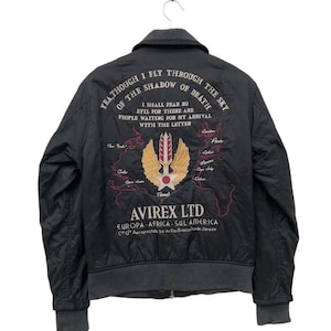 Puede incluir: Chaqueta bomber negra con texto bordado y dise&ntilde;o de mapa. La chaqueta tiene cuello, pu&ntilde;os y dobladillo grises. La parte trasera muestra el texto "YEA THOUGH I FLY THROUGH THE SKY OF THE SHADOW OF DEATH" y un logotipo con el texto "AVIREX LTD".