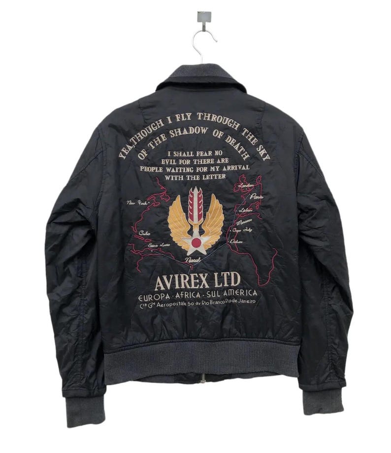Puede incluir: Chaqueta bomber negra con texto bordado y dise&ntilde;o de mapa. La chaqueta tiene cuello, pu&ntilde;os y dobladillo grises. La parte trasera muestra el texto "YEA THOUGH I FLY THROUGH THE SKY OF THE SHADOW OF DEATH" y un logotipo con el texto "AVIREX LTD".