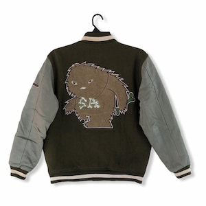 Peut inclure: Une veste de baseball verte et grise avec un motif de monstre brun au dos. Le monstre porte un collier avec des os et un nœud papillon vert. La veste a une bande blanche autour du col et des poignets.