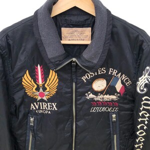 Puede incluir: Chaqueta bomber acolchada negra con parches bordados. Presenta un emblema dorado y rojo, el texto "AVIREX EUROPA" y "POSTES FRANCE" con una bandera francesa. Incluye cuello gris y dos bolsillos con cremallera.