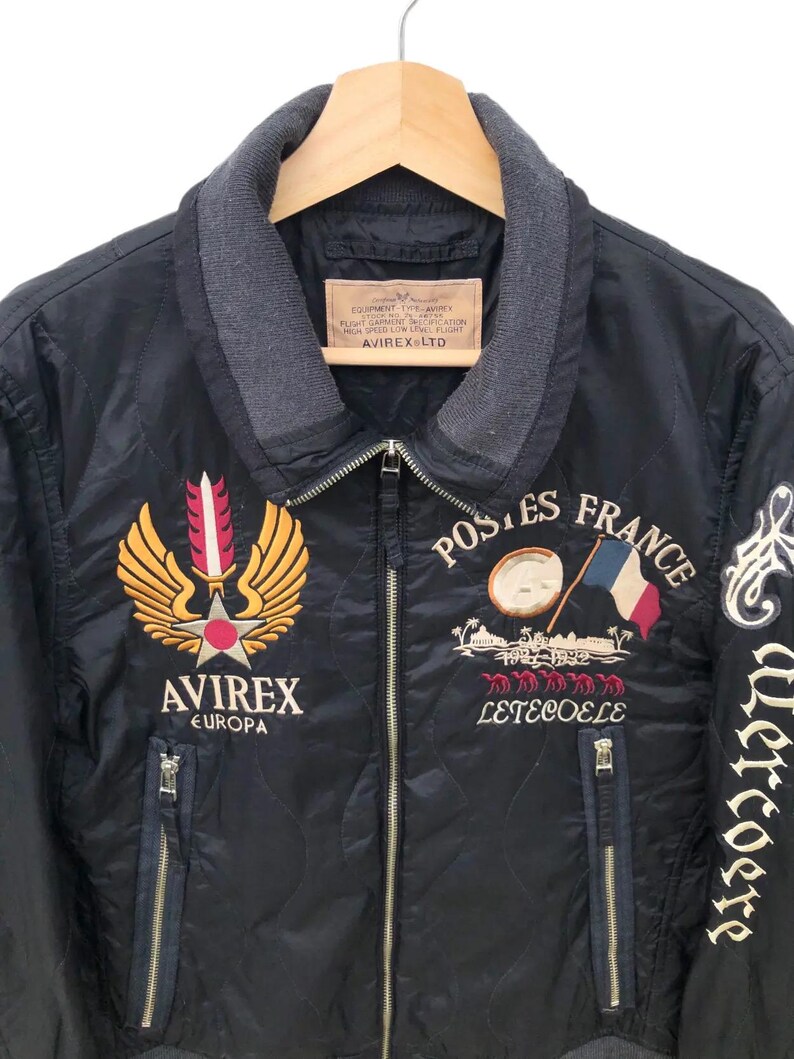 Puede incluir: Chaqueta bomber acolchada negra con parches bordados. Presenta un emblema dorado y rojo, el texto "AVIREX EUROPA" y "POSTES FRANCE" con una bandera francesa. Incluye cuello gris y dos bolsillos con cremallera.