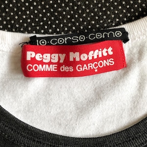 Puede incluir: Etiqueta roja y blanca con el texto "Peggy Moffitt COMME des GARCONS" en una camiseta blanca con cuello negro.