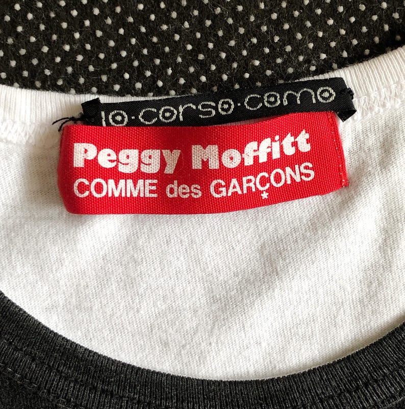 Puede incluir: Etiqueta roja y blanca con el texto "Peggy Moffitt COMME des GARCONS" en una camiseta blanca con cuello negro.