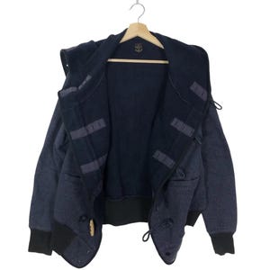 Può includere: Una felpa con cappuccio blu scuro con chiusura a zip e chiusura a bottone. La felpa ha un design unico con un cappuccio a strati e una fodera nera a contrasto.