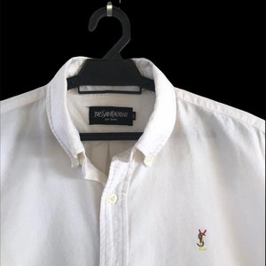 Puede incluir: Camisa blanca de manga larga con botones y un pequeño logotipo YSL bordado en el pecho. La camisa tiene una etiqueta negra con el texto "Yves Saint Laurent pour Homme" en el cuello.