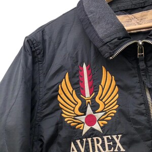 Puede incluir: Chaqueta bomber negra con cuello gris y cierre de cremallera. La chaqueta presenta un emblema bordado con alas doradas, una estrella blanca y un c&iacute;rculo rojo. El texto "AVIREX EUROPA" est&aacute; bordado debajo.