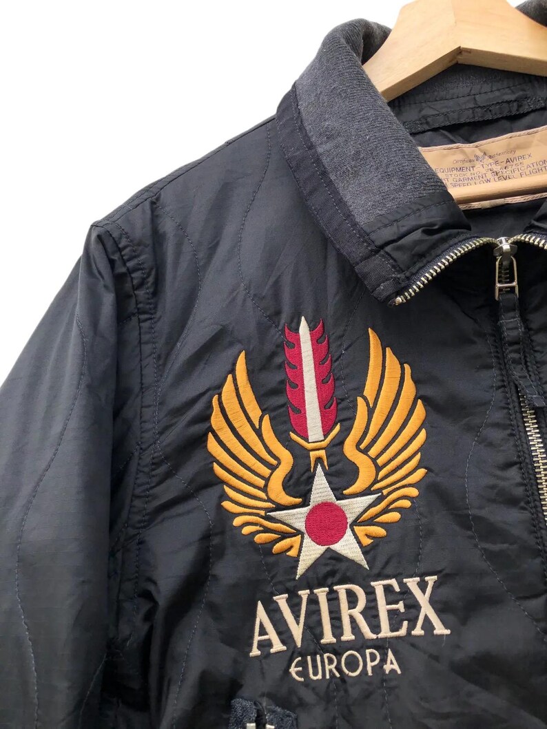 Puede incluir: Chaqueta bomber negra con cuello gris y cierre de cremallera. La chaqueta presenta un emblema bordado con alas doradas, una estrella blanca y un c&iacute;rculo rojo. El texto "AVIREX EUROPA" est&aacute; bordado debajo.