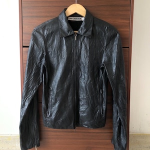 Peut inclure: Veste en cuir noire avec fermeture éclair et col. La veste est suspendue à un cintre en bois. Le nom de la marque "JIL SANDER" est visible sur l'étiquette.