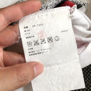 Puede incluir: Etiqueta de ropa blanca con texto negro que dice "ZK-T003", "Algod&oacute;n 100%", "Talla", "Hecho en Jap&oacute;n", "Commes des Gar&ccedil;ons", y "Tokio, Jap&oacute;n". La etiqueta tambi&eacute;n incluye un gr&aacute;fico en blanco y negro de un s&iacute;mbolo de instrucciones de lavado.