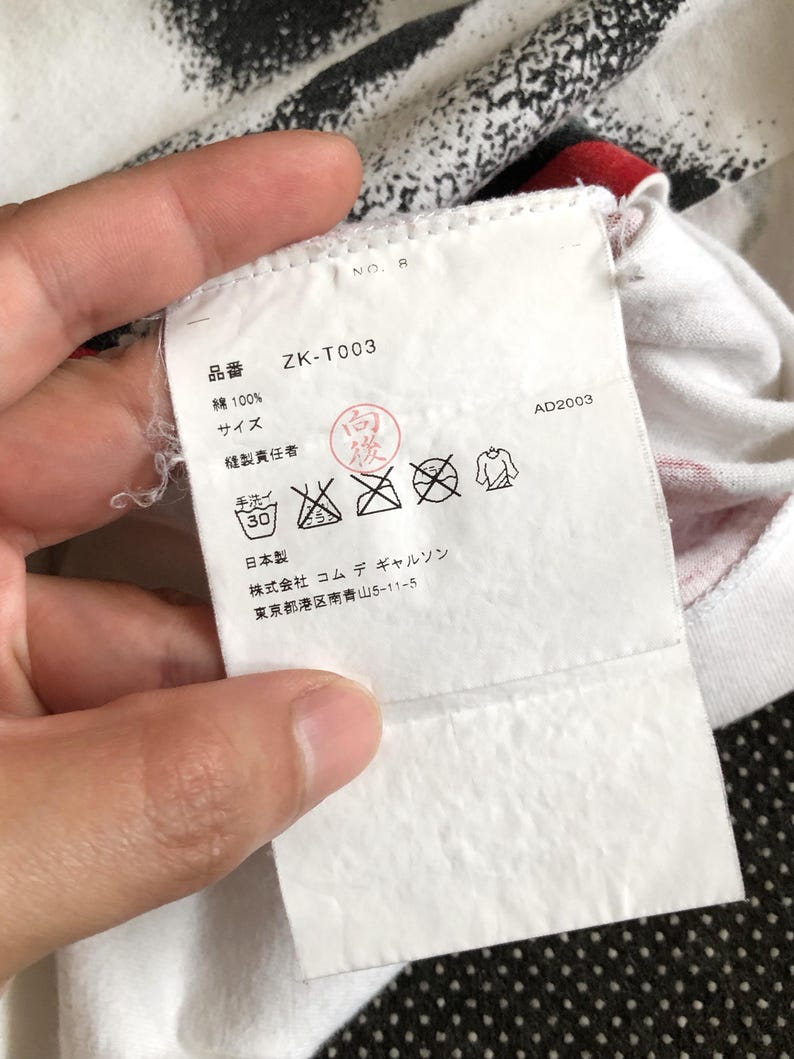 Puede incluir: Etiqueta de ropa blanca con texto negro que dice "ZK-T003", "Algod&oacute;n 100%", "Talla", "Hecho en Jap&oacute;n", "Commes des Gar&ccedil;ons", y "Tokio, Jap&oacute;n". La etiqueta tambi&eacute;n incluye un gr&aacute;fico en blanco y negro de un s&iacute;mbolo de instrucciones de lavado.