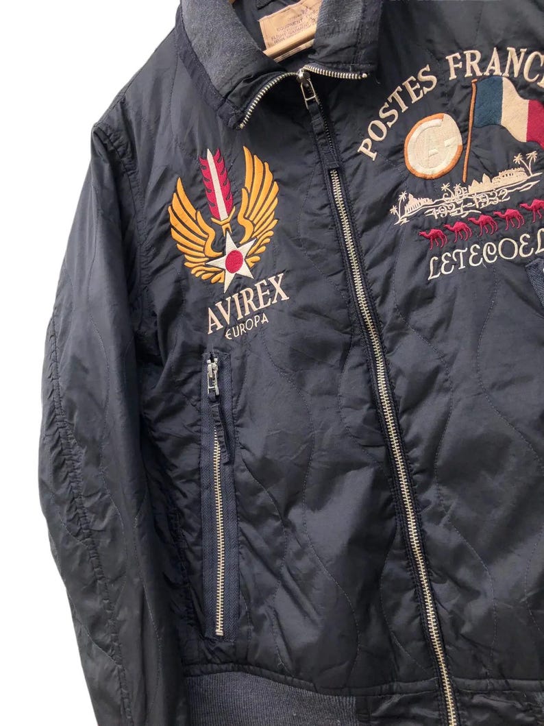 Puede incluir: Chaqueta acolchada azul marino con parches bordados. Presenta los textos "POSTES FRANCE" y "AVIREX EUROPA". Incluye cierre de cremallera y cuello gris. La chaqueta tiene un patr&oacute;n de diamantes.