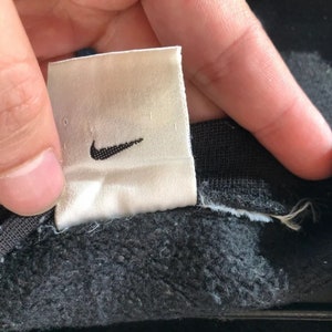 May include: A white tag with a black Nike swoosh logo. The tag is attached to a black garment.