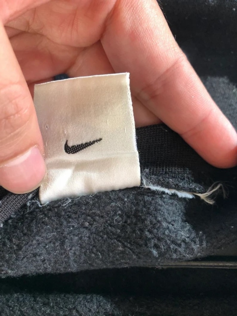 May include: A white tag with a black Nike swoosh logo. The tag is attached to a black garment.