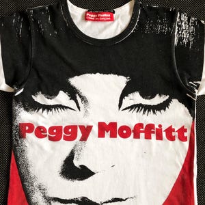 Puede incluir: Camiseta gr&aacute;fica en blanco y negro con una imagen roja y blanca de la cara de una mujer y el texto "Peggy Moffitt" en rojo. La camiseta tiene una etiqueta roja con el texto "Peggy Moffitt Comme des Gar&ccedil;ons" en la parte delantera.