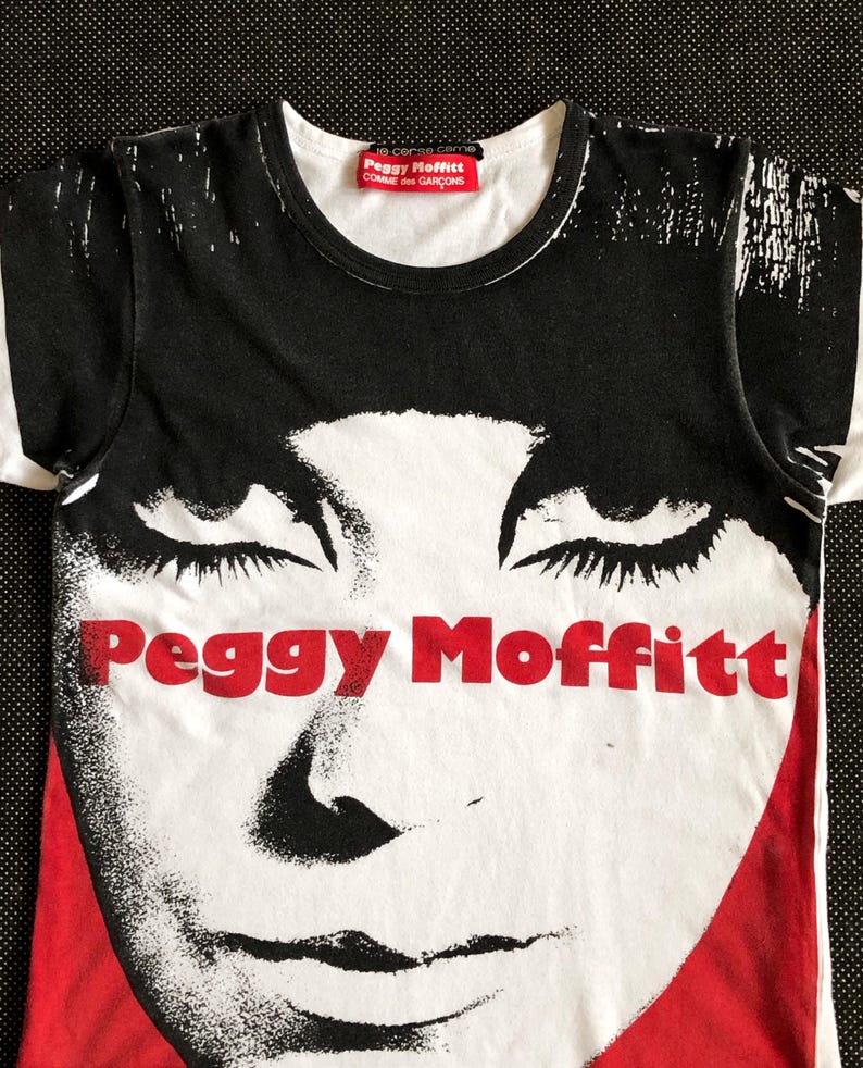 Puede incluir: Camiseta gr&aacute;fica en blanco y negro con una imagen roja y blanca de la cara de una mujer y el texto "Peggy Moffitt" en rojo. La camiseta tiene una etiqueta roja con el texto "Peggy Moffitt Comme des Gar&ccedil;ons" en la parte delantera.