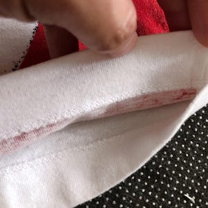 Puede incluir: Primer plano del interior de la costura de una camiseta blanca con una franja roja a lo largo del borde.
