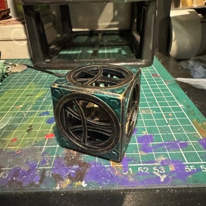 May include: A small, cube-shaped decorative object with a geometric design. The cube is made of wood and painted in shades of green and black.