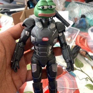 Op de afbeelding: Een zwart en zilveren action figure van Iron Man met een groene kikkerkop. De figuur houdt een zwart wapen in zijn rechterhand.