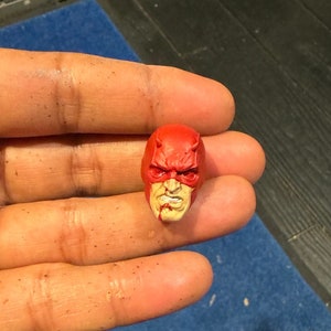 Könnte beinhalten: Ein roter und weißer bemalter Plastik-Actionfigur-Kopf von Daredevil, einem Superhelden mit einer roten Maske und einem weißen Gesicht. Die Figur hat ein detailliertes Gesicht mit einer gerunzelten Stirn, einem entschlossenen Ausdruck und einer blutigen Nase.