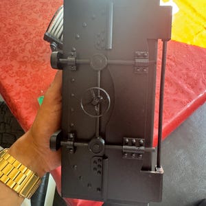 Puede incluir: Una caja negra rectangular con una puerta con bisagras, que se asemeja a una caja fuerte. La caja presenta un dial circular, un asa en forma de estrella y múltiples remaches. El diseño general es industrial y detallado, con un acabado mate.