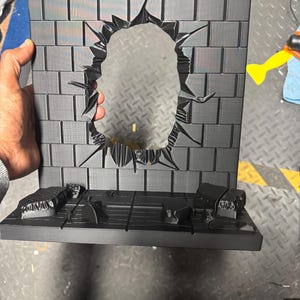 Może przedstawiać: Czarna, drukowana w 3D podstawa dioramy przedstawiająca ceglany mur z otworem pośrodku, jakby coś się przez niego przebiło. Podstawa zawiera platformę z dodatkowymi rzeźbionymi detalami, idealną do eksponowania figurek akcji lub przedmiotów kolekcjonerskich.