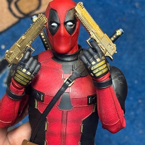 May include: An action figure of Deadpool, dressed in a red and black suit, holding two gold-coloured pistols. The figure has a brown belt and a black strap across the chest.