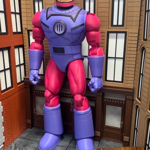 May include: A large action figure of a Sentinel robot with a pink helmet, purple chest armor, and pink limbs. The figure features a circular chest emblem with three vertical lines, articulated joints, and a blocky, mechanical design standing upright.