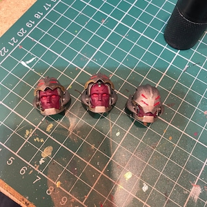 May include: Three silver and red plastic helmets with red eyes. The helmets are on a green cutting mat.