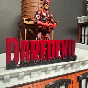 Op de afbeelding: Een rood en zwart Daredevil actiefiguur staat op een dakset, met een watertoren op de achtergrond. Het woord "DAREDEVIL" wordt weergegeven in rode letters met een zwarte skyline basis. Het gebouw is gemaakt van rode bakstenen.