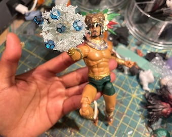 May include: A plastic action figure of a man wearing a gold and green costume, holding a clear plastic object with blue gems. The figure is standing on a green and white checkered surface.