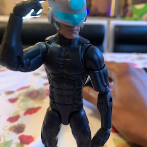 May include: A black action figure with a silver helmet and a blue visor. The figure is posed with one hand on its head and the other arm bent at the elbow.