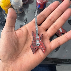 Peut inclure: Une guitare électrique miniature rouge avec des accents noirs et une touche blanche. La guitare est tenue dans la main d'une personne.