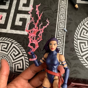 May include: A blue and black action figure of a female superhero with purple hair and a pink lightning bolt accessory. The figure is posed with one arm raised and the other holding the accessory.