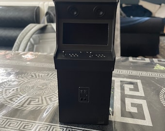 May include: A black miniature arcade cabinet with a small screen, buttons, and a joystick. The cabinet is sitting on a table with a patterned tablecloth.