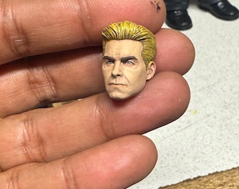 May include: A detailed, hand-painted miniature head with blonde hair and a stern expression. The figure's face is painted with realistic skin tones and features, showcasing fine details. The head is held in a person's hand.