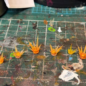 Puede incluir: Colección de efectos de llama en miniatura, translúcidos, de color naranja y amarillo, probablemente para modelismo o juegos de miniaturas. Dispuestos sobre una superficie de cuadrícula verde, sugiriendo un entorno de manualidades.