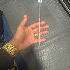 May include: A silver and gold spear with a triangular blade. The spear has gold accents and a silver shaft. The spear is held in a hand, with a gold watch visible on the wrist. The background is a textured grey surface.