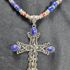 May include: A silver necklace with a cross pendant featuring blue gemstone accents. The necklace is made of silver beads and red and pink beads.