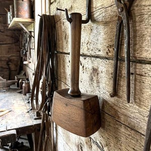May include: A dark brown wooden mallet with a long, cylindrical handle and a wide, rectangular head featuring a visible wood grain pattern. The mallet is suspended from a metal hook attached to a rough, horizontal wooden plank wall.