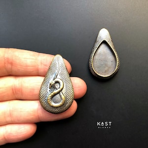 Puede incluir: Dos piezas en blanco de metal en forma de lágrima para la elaboración de joyas. Una pieza en blanco presenta un diseño de serpiente en relieve con un acabado dorado. La otra pieza en blanco es lisa y tiene un acabado plateado. KAST BLANKS está impreso en el fondo.
