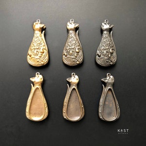 May include: Six metal cat-shaped pendant blanks in three finishes: gold, brass, and black. The blanks are detailed with floral designs and have a smooth, oval-shaped interior.