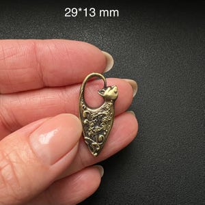 Op de afbeelding: Een bronzen katvormige hanger met ingewikkelde bloemen- en wervelontwerpen. De hanger is 2,9 cm lang en 1,3 cm breed. De kattenkop vormt de bovenste lus, en het lichaam loopt taps toe.