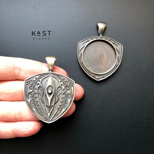 Könnte beinhalten: Zwei bronzene Anhänger in Schildform. Ein Anhänger hat ein detailliertes Design einer stilisierten Feder mit einem kleinen Kreis in der Mitte. Der andere Anhänger ist schlicht mit einer runden Öffnung in der Mitte.