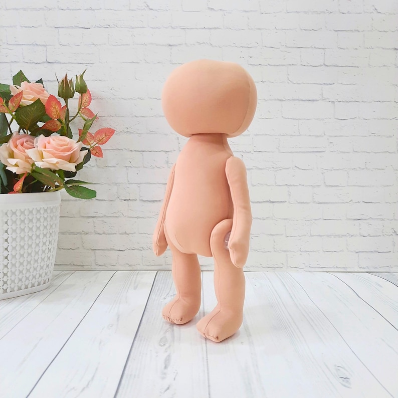 May include: A plain, unpainted doll body made of fabric. The doll is standing upright with its arms at its sides. The doll is peach colored.