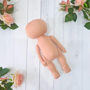 May include: A nude-colored fabric doll with a round head and no facial features. The doll is lying on a white wooden surface.