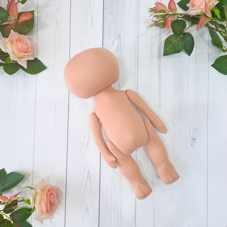 May include: A nude-colored fabric doll with a round head and no facial features. The doll is lying on a white wooden surface.