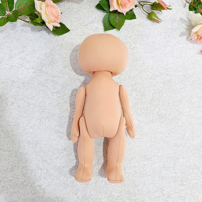 May include: A nude-colored fabric doll with a round head and no facial features. The doll is posed standing with its arms at its sides and its legs slightly apart.