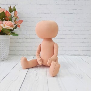 May include: A nude-colored fabric doll with a large head and limbs. The doll is sitting with its legs crossed and its arms folded in front of it.